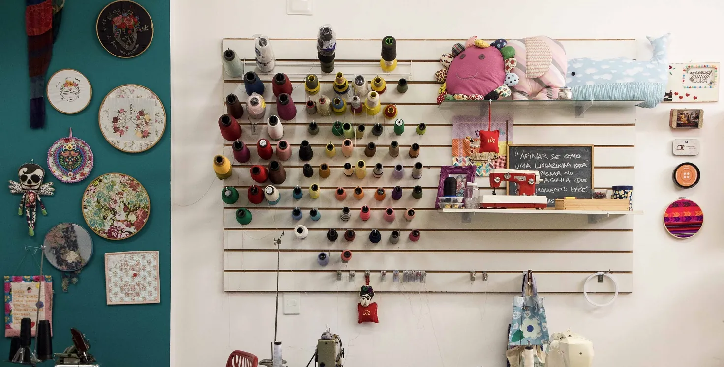 Vista do ateliê: bastidores de bordado e quadros de tecido pendurados na parede verde-azulada